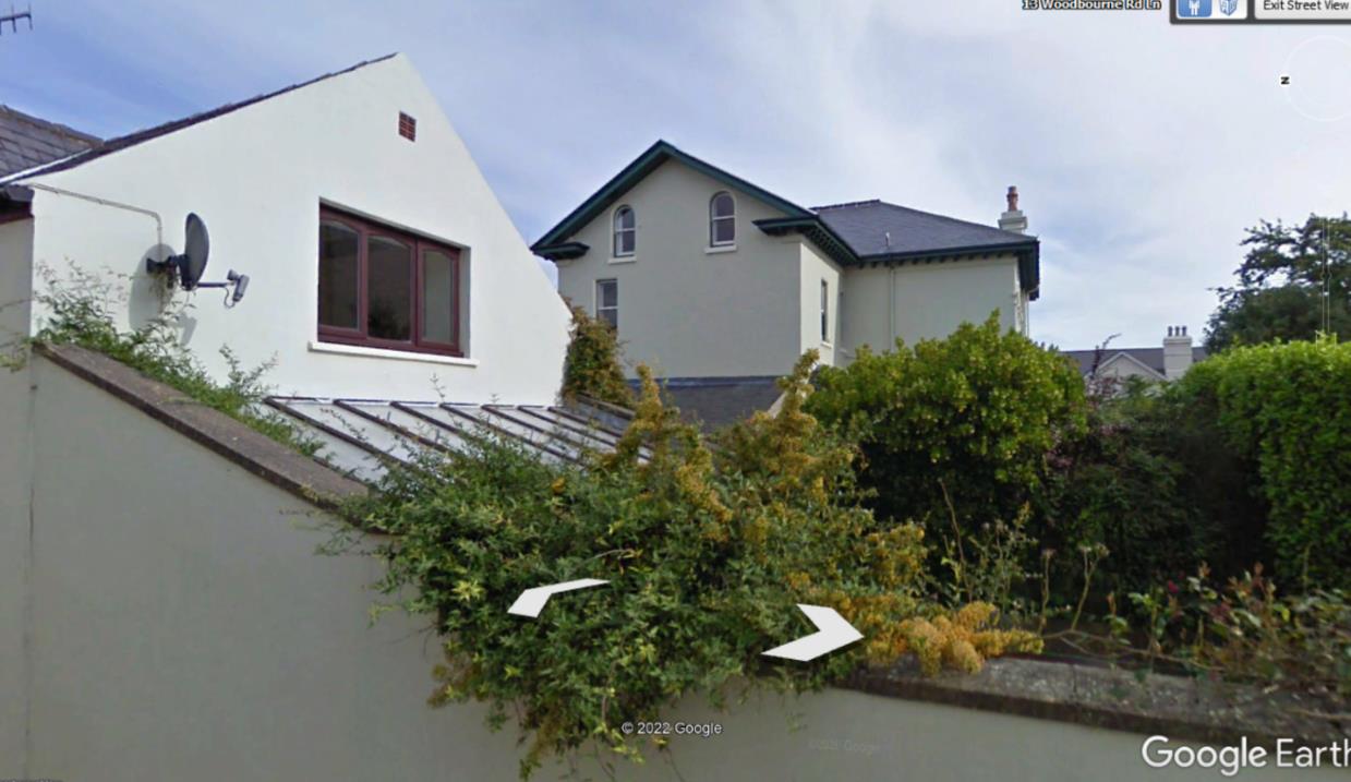 A Google Earth screenshot showing a white building and a cream detached house, with white arrows overlaid pointing towards foreground vegetation and a boundary wall.