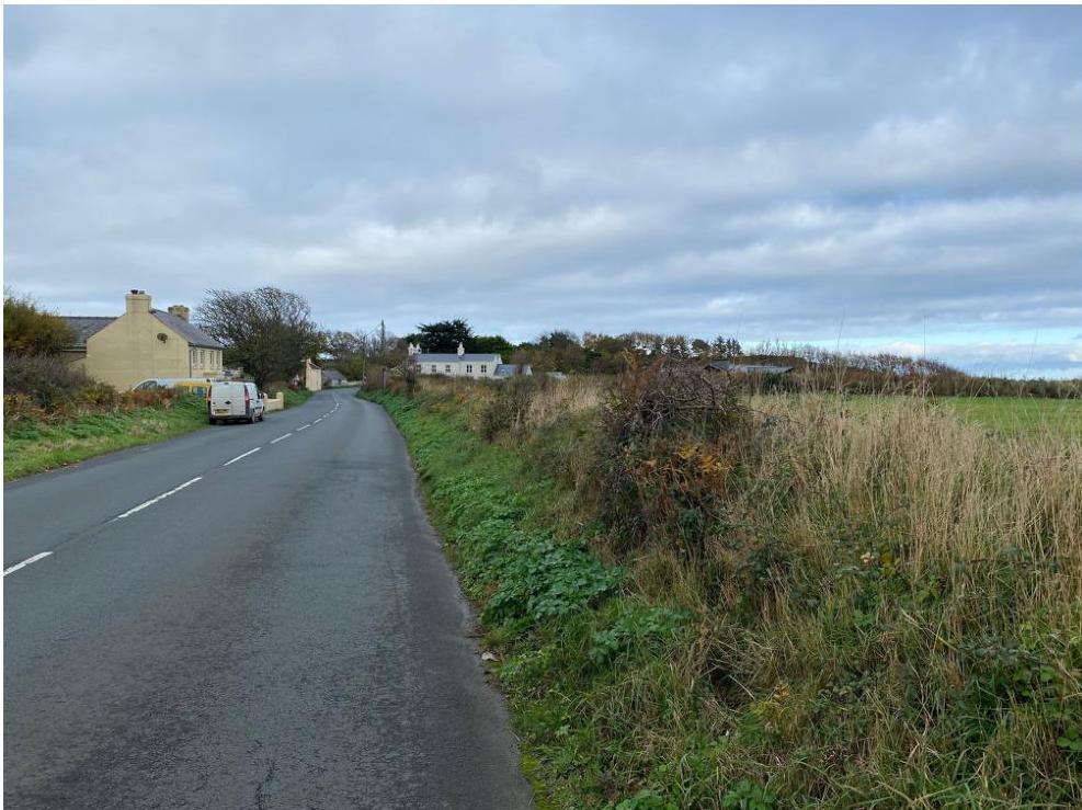 A photograph showing a rural road scene with existing houses on the left and a grassy field on the right.