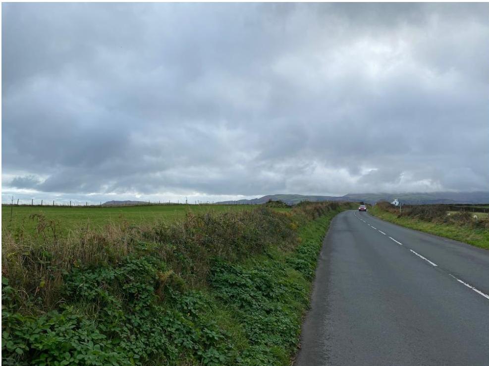 A photograph showing a paved road curving through green agricultural fields under a cloudy sky with hills in the background.