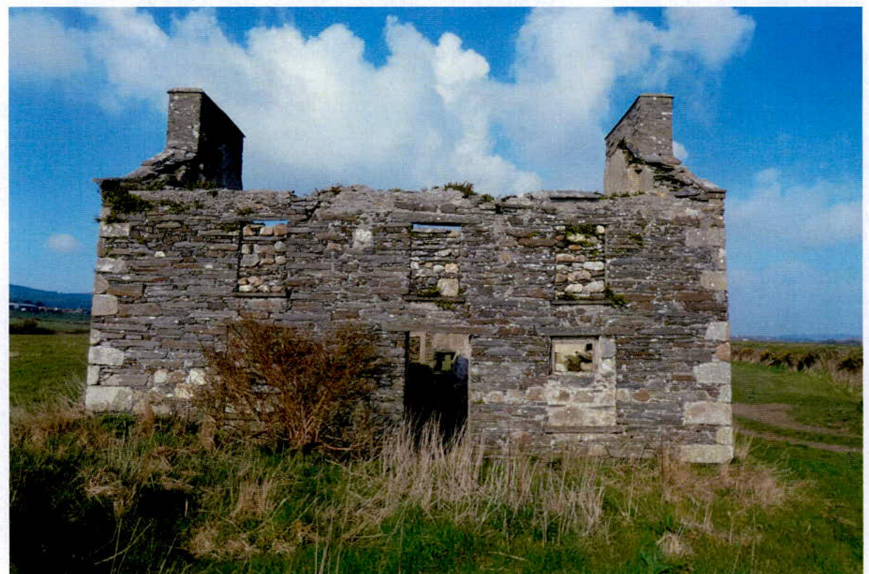 A photograph showing the exterior of a dilapidated stone ruin with two chimneys and no roof, set in a grassy rural landscape.