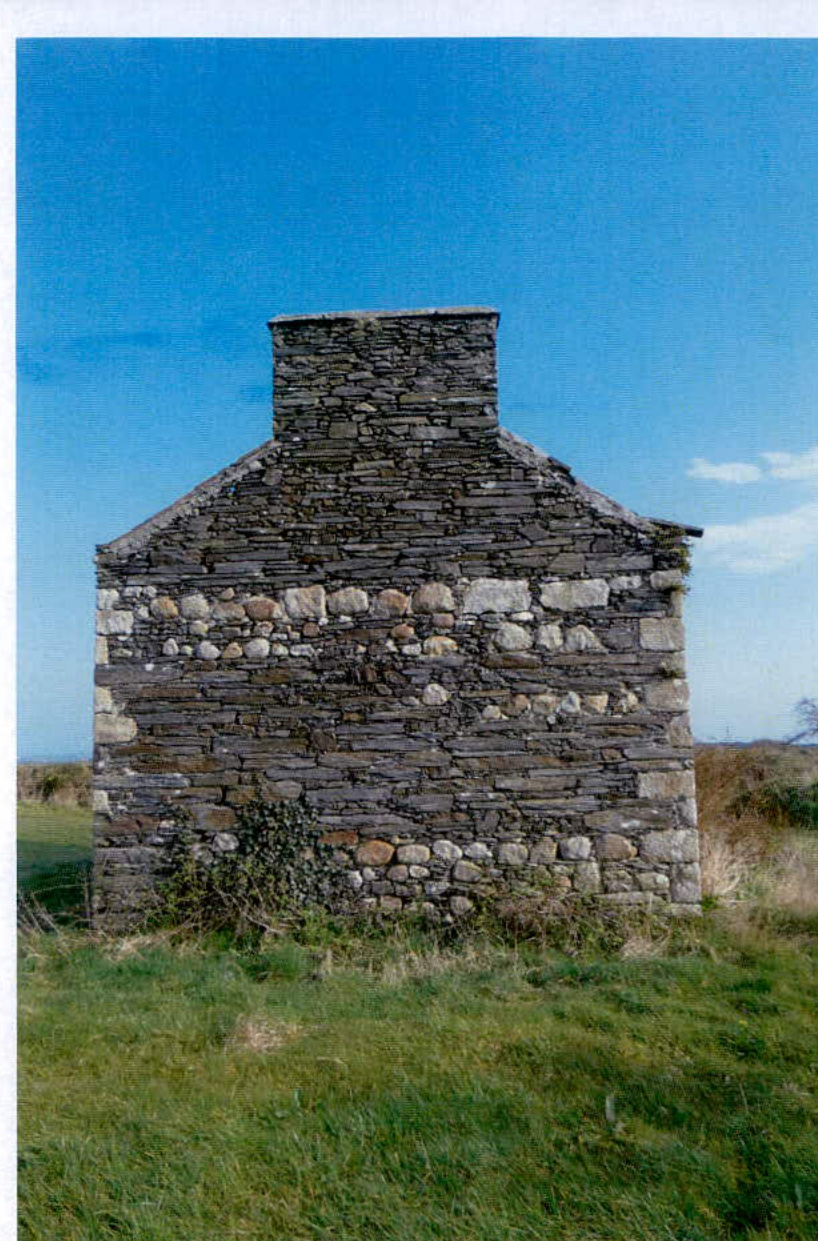 A photograph showing the stone gable end of a building, likely a barn or cottage, standing in a grassy field under a blue sky.