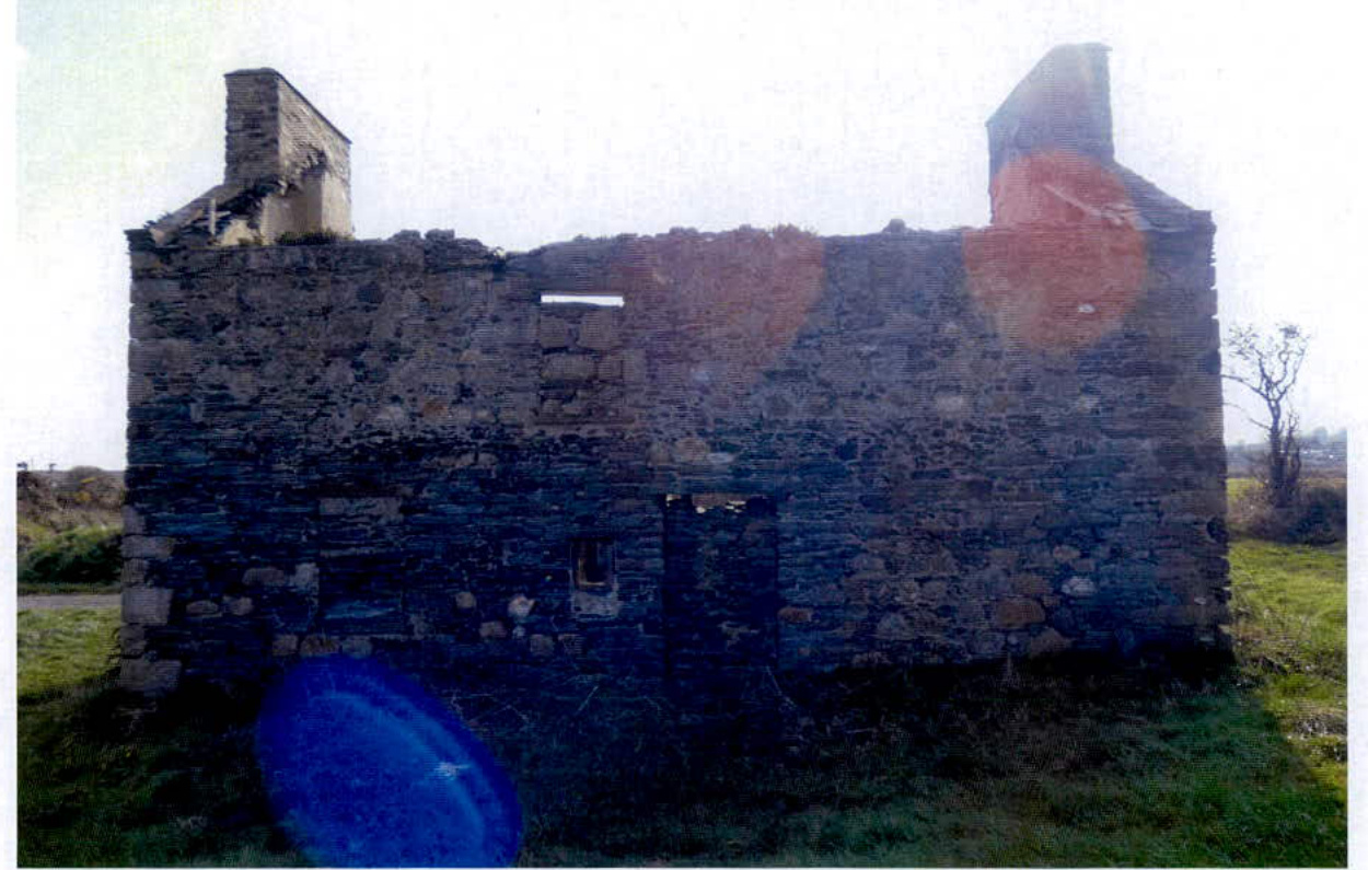 A photograph showing the exterior of a derelict stone cottage ruin with two chimneys and missing roof, situated in a grassy rural setting.