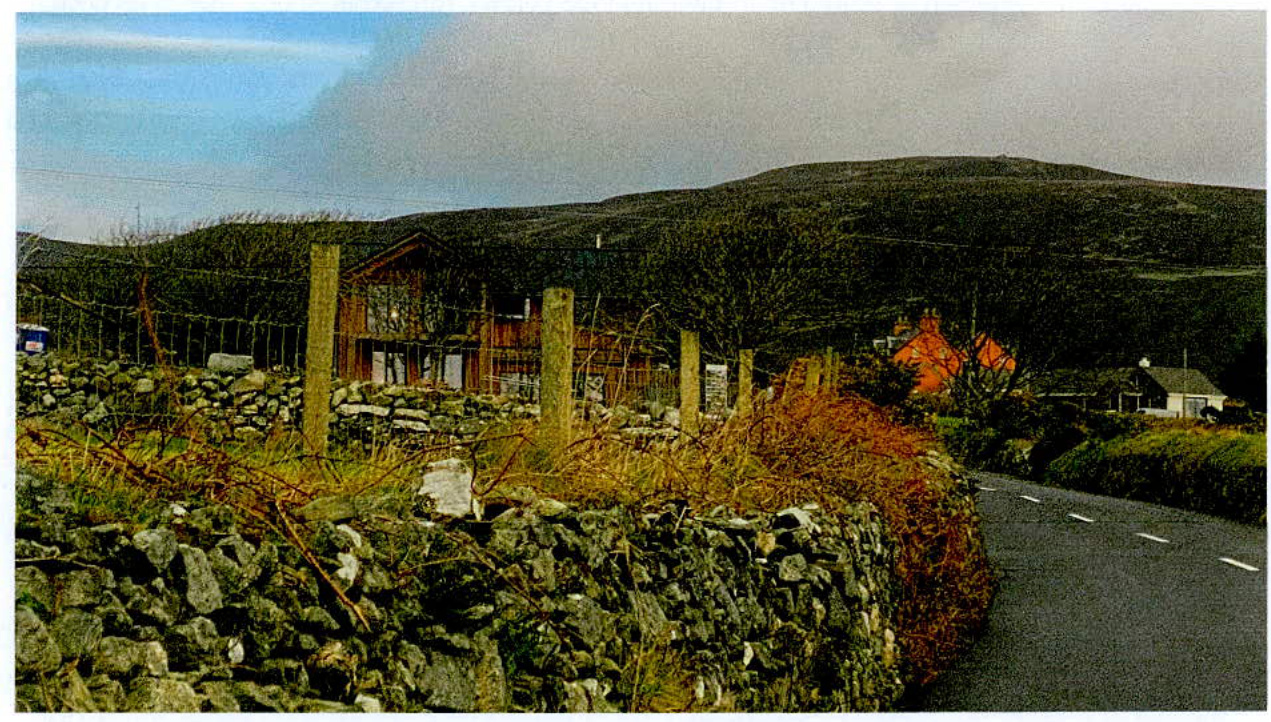 A photograph of a rural site featuring a wooden agricultural building next to a road and dry stone walls, with hills in the background.