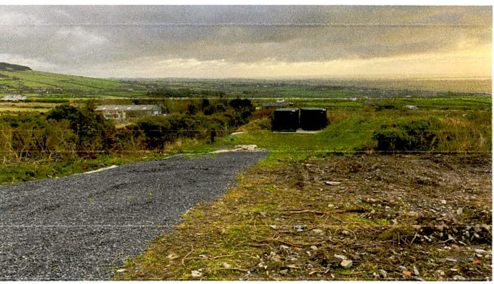 A photograph of a rural site showing a gravel driveway and two dark rectangular structures, likely the proposed pump house, with a coastal view in the distance.
