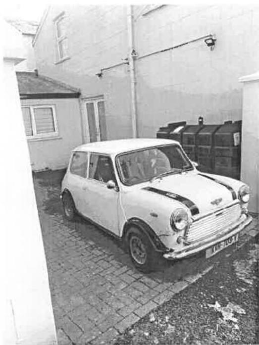 A black and white photograph showing a white classic Mini car parked on a paved driveway next to a building and large waste bins.