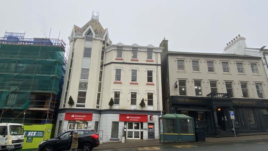 A street-level photograph showing a row of commercial buildings including a Santander bank branch and a pub, with a building under construction covered in scaffolding to the left.