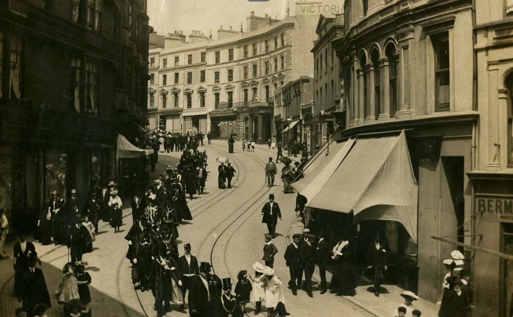 A vintage sepia-toned photograph depicting a busy street scene with pedestrians in period clothing, horse-drawn carriages, and tram tracks.