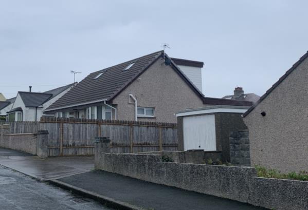 A street-level photograph showing the rear elevation of a detached house with a pitched roof and skylights, viewed across a wooden fence and boundary wall.