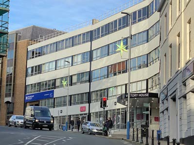 A street-level photograph of a multi-story commercial building identified as Victory House, showing the existing facade, street, and parked vehicles.