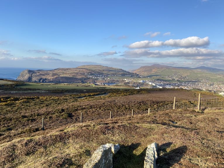 A wide landscape photograph showing a coastal town in the distance with rolling hills and rough terrain in the foreground.