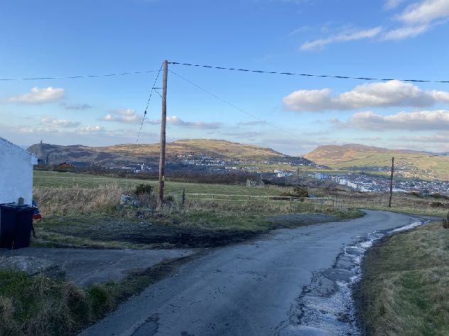 A photograph showing a rural road curving through grassy fields with hills and a town visible in the background.