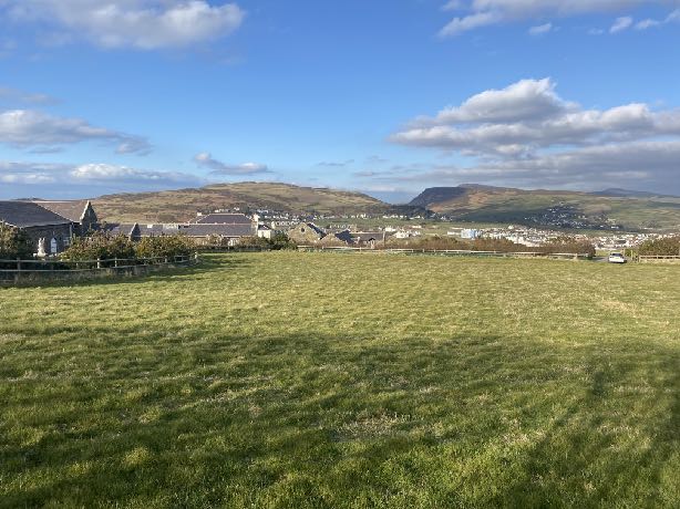 A landscape photograph showing a large grassy field in the foreground with a wooden fence, overlooking a village and hills in the distance.