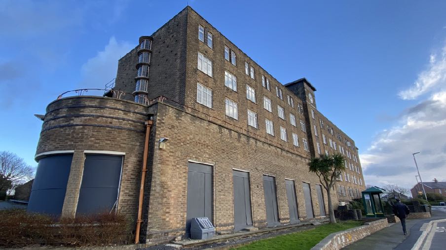 A low-angle exterior photograph of a large, multi-story brick building featuring a distinctive curved corner section and ground-floor garage-style doors. The building has numerous white-framed windows and is situated...