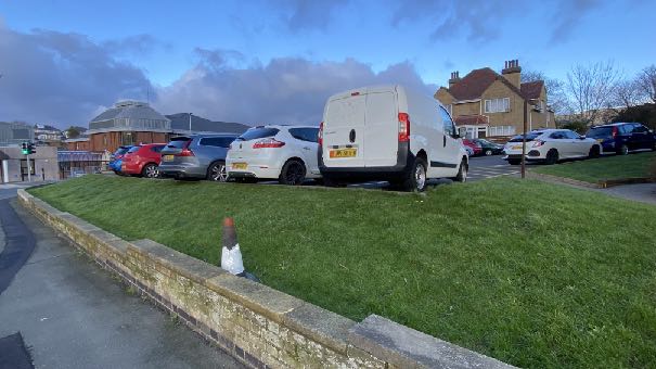 A photograph showing a grassy verge with a stone wall in the foreground and a parking area with cars behind it. Background buildings include a brick house and a structure with a glass roof.