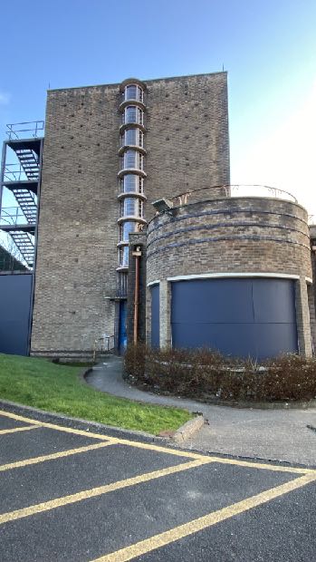 A low-angle photograph of a tall brick building featuring a rectangular tower section with a vertical window strip and an adjacent cylindrical brick structure. An external metal staircase is visible on the left, and a...