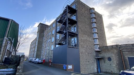 A street-level photograph showing a large multi-story brick building with a prominent external metal staircase and a new dark grey extension at the base.