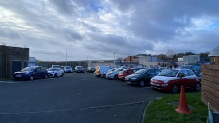 A photograph showing a paved car park with several parked vehicles, a grass verge with an orange cone in the foreground, and various buildings in the background under a cloudy sky.