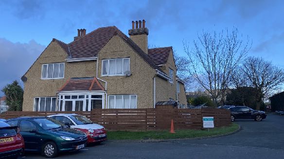 A photograph of a large, two-story residential building with pebbledash walls and a tiled roof. Several cars are parked in front of a wooden fence with a planning application notice attached.