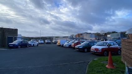 A photograph showing a paved parking area with several cars parked in rows, featuring a brick wall in the foreground and commercial-style buildings in the background.