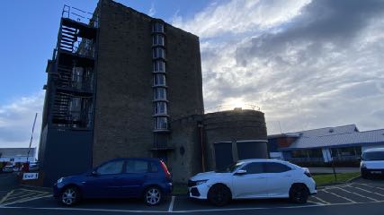 A photograph of a tall brick building with a modern dark grey extension featuring balconies. Cars are parked in a lot in the foreground under a cloudy sky.