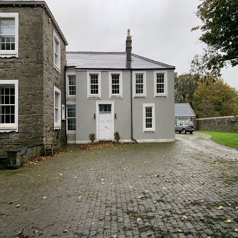 A photograph showing the exterior of a building with a stone wing and a grey rendered wing, viewed from a paved driveway.