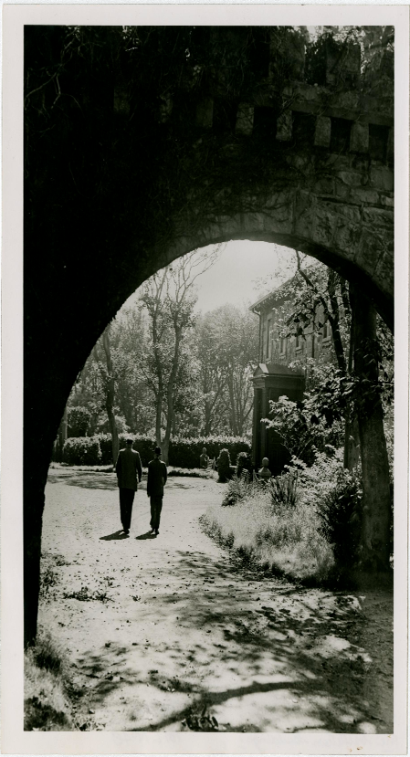 A black and white photograph showing a stone archway framing a view of a driveway with two people walking towards a house in the background.