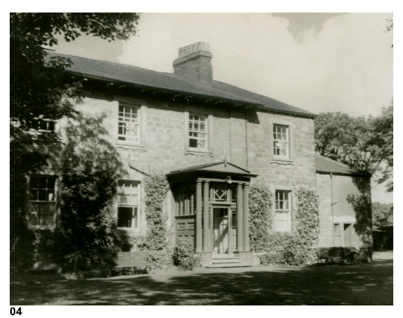 A black and white photograph showing the exterior of a large, two-story stone house with a central portico entrance and ivy-covered walls.