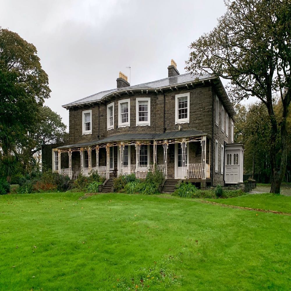 A photograph of a large, two-story stone house featuring a prominent white veranda with intricate ironwork. The property is set within a spacious green lawn surrounded by mature trees.