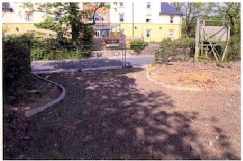 A photograph showing a gravel driveway and cleared land in the foreground with a large yellow building in the background.