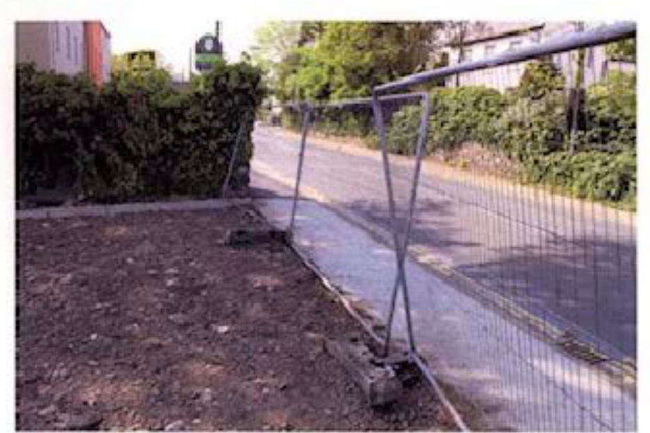 A photograph showing a vacant plot of land with bare earth, fenced off from the street with temporary metal barriers.
