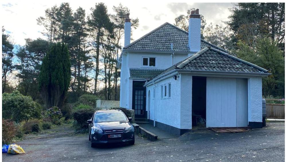 A photograph showing the exterior of a white two-story detached house with an attached single-story garage or extension, set in a rural environment with trees.