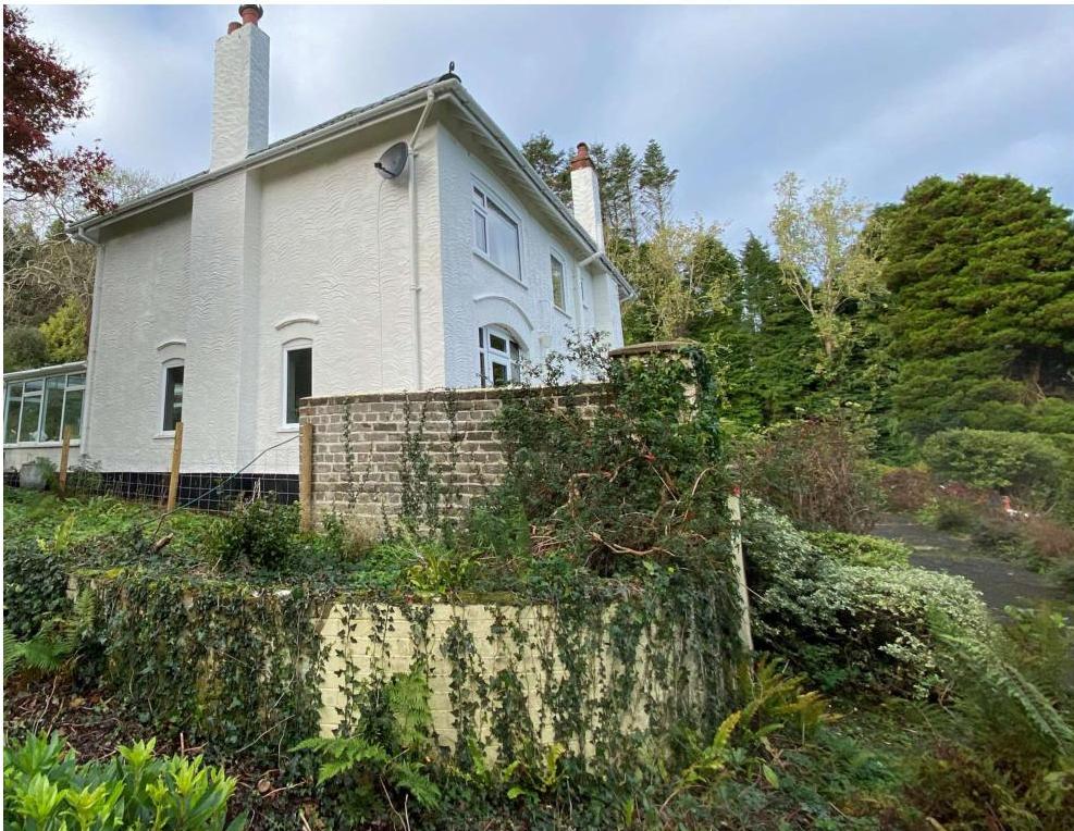 A photograph showing a white, two-story detached house with a glass conservatory extension on the left. The property features brick retaining walls and is surrounded by trees and vegetation.
