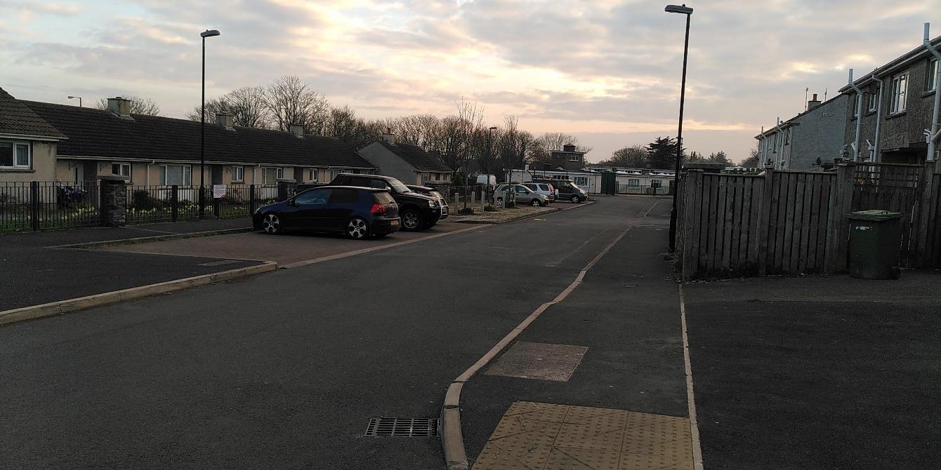 A street-level photograph showing a paved road with parked cars, terraced bungalows on the left, and a wooden fence on the right.