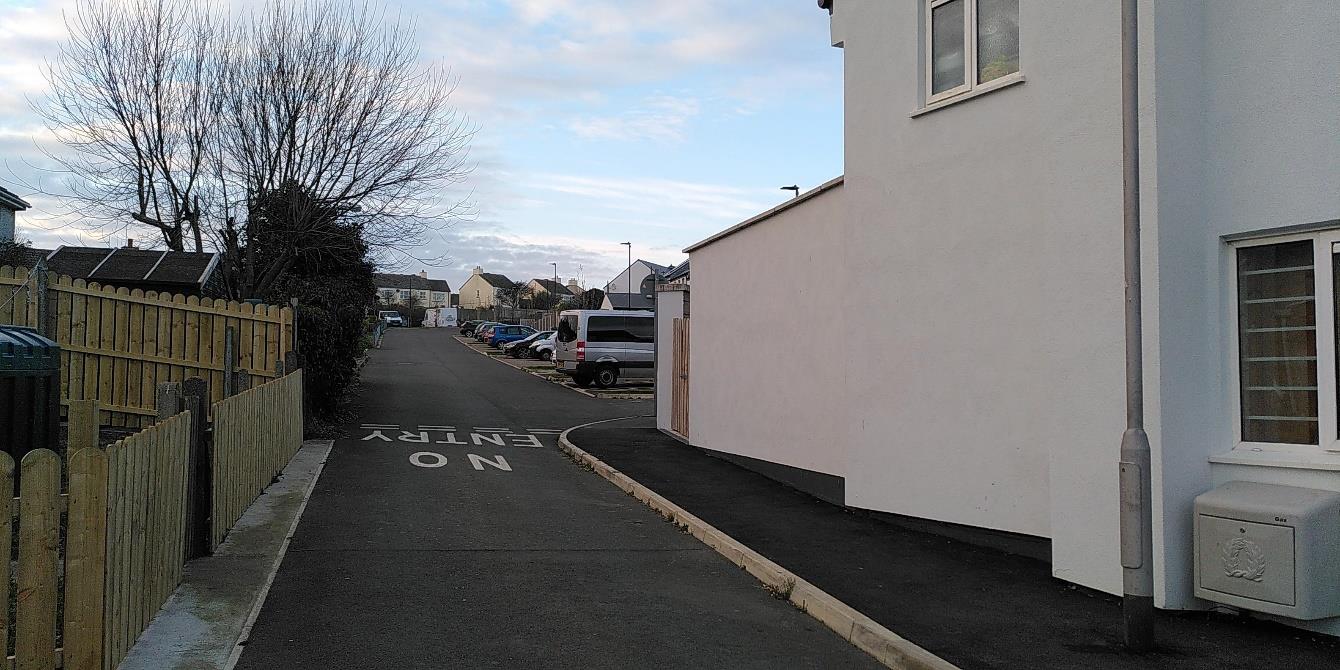 A street-level photograph showing a white building on the right and a wooden fence on the left, with a paved road marked 'NO ENTRY' leading towards a parking area in the background.
