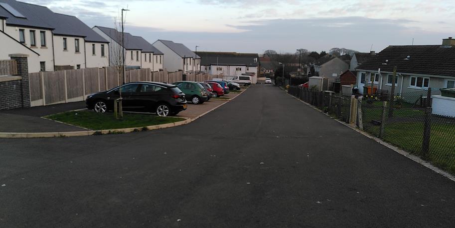 A street-level photograph showing a residential road with modern white houses and parking bays on the left, and older bungalows on the right.