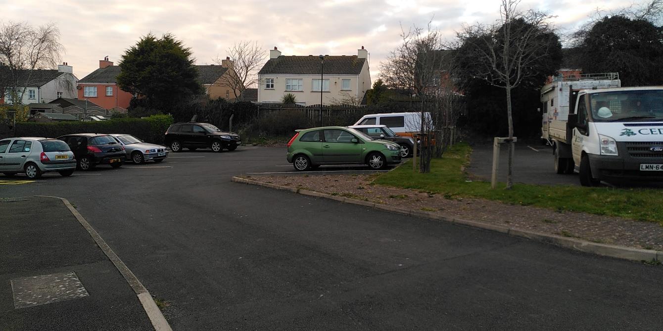 A street-level photograph showing a paved parking area with several cars and a white van, with residential houses visible in the background.