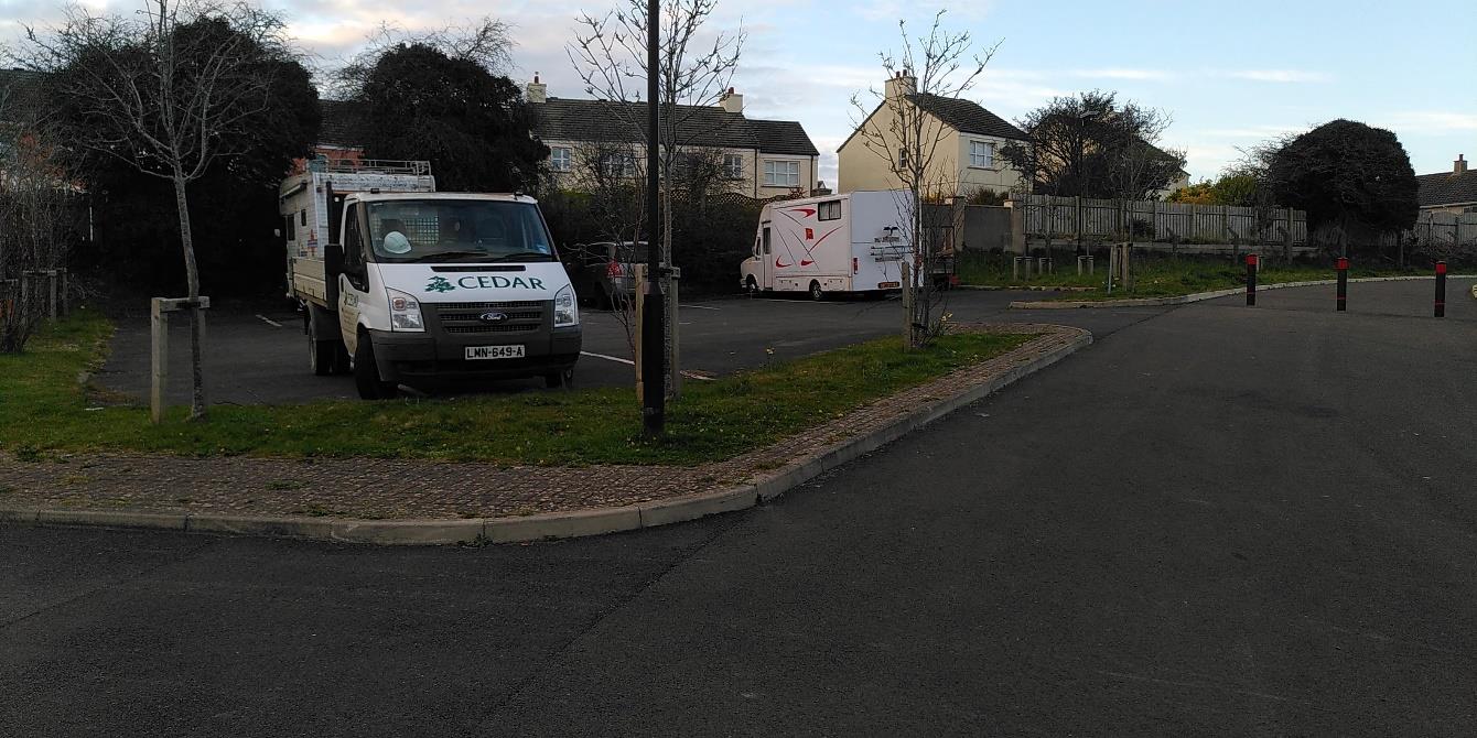 A photograph showing a paved parking area with a work truck and a camper van, with residential houses and trees visible in the background.