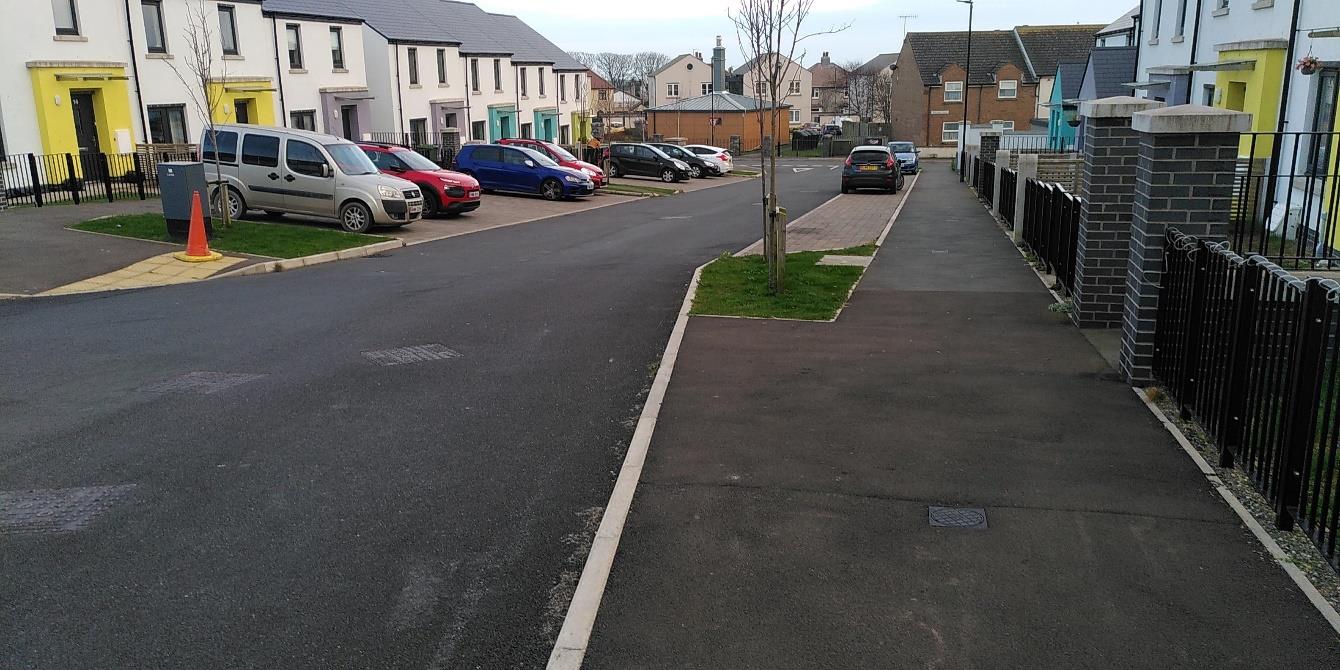 A street-level photograph showing a row of terraced houses with cars parked along the curb and a paved sidewalk on the right.