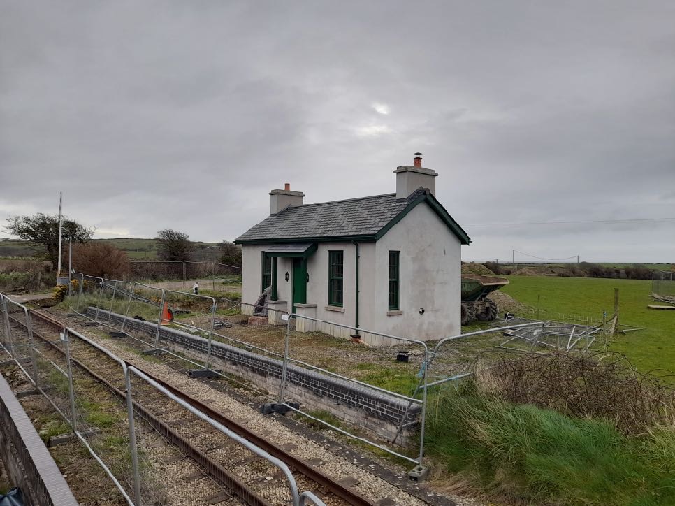 A photograph showing a small white single-story cottage with green trim situated next to a railway track in a rural setting.