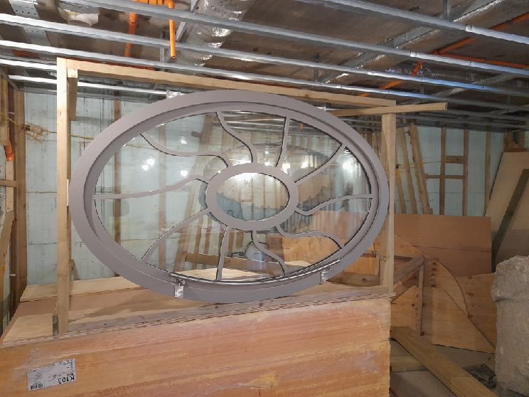 A large, oval-shaped decorative window with a metal grille design resting on a wooden crate within an unfinished interior space.