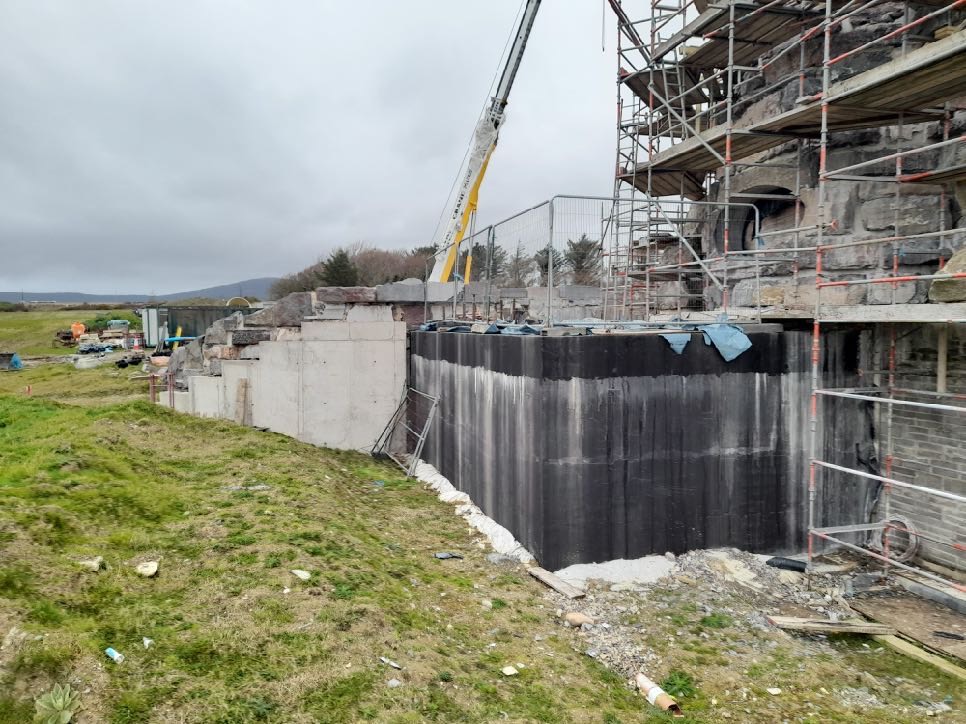 A photograph showing a construction site with a stone building covered in scaffolding and a large retaining wall with black waterproofing membrane.