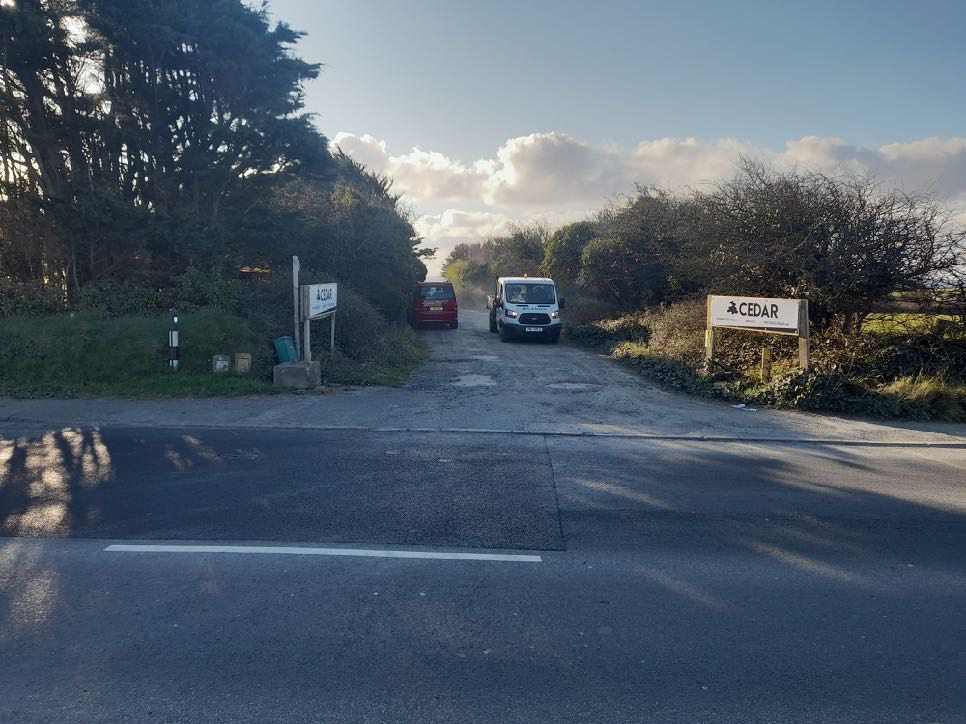 A photograph showing the entrance to a property named 'Cedar' with vehicles parked inside, viewed from the roadside.