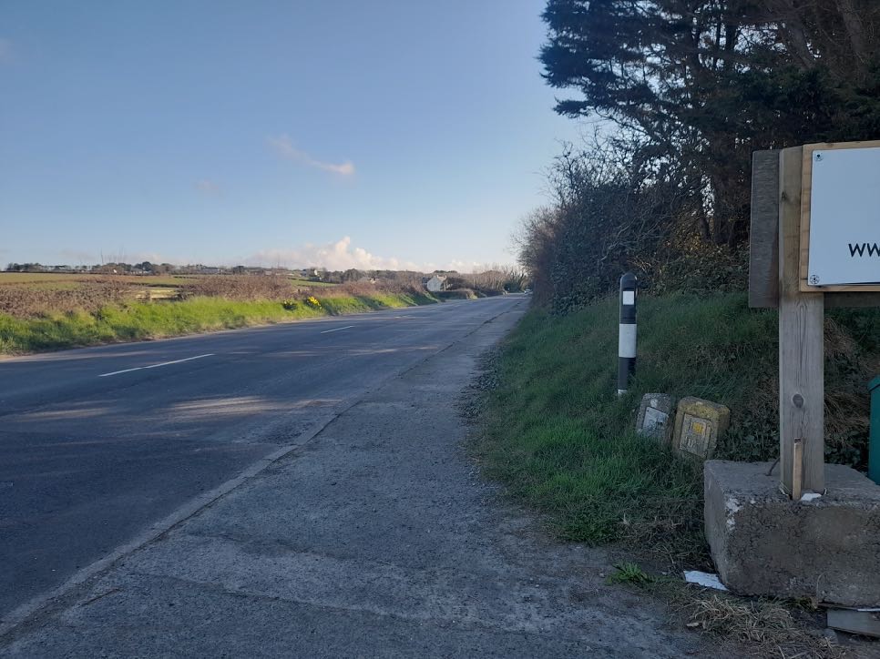 A photograph showing a rural road scene with a grassy verge, a black and white bollard, and a wooden signpost on the right side.