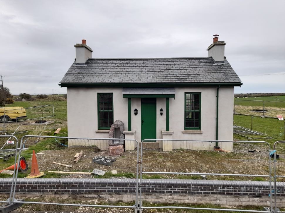 A photograph showing the exterior of a white cottage with green window frames and a slate roof, surrounded by construction fencing and unfinished ground.