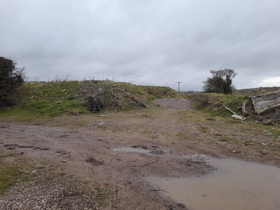 A muddy, unpaved access track or entrance area with grassy embankments on either side under an overcast sky.