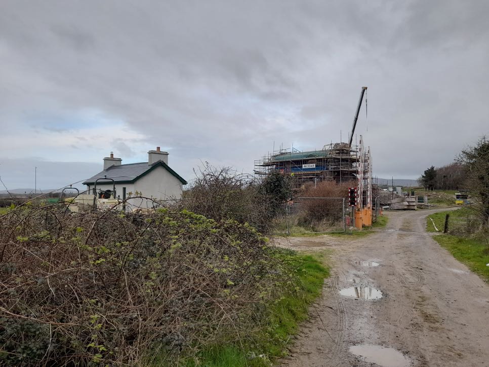 A photograph showing a white detached cottage on the left and a large building under construction with scaffolding and a crane on the right, viewed from a muddy track.