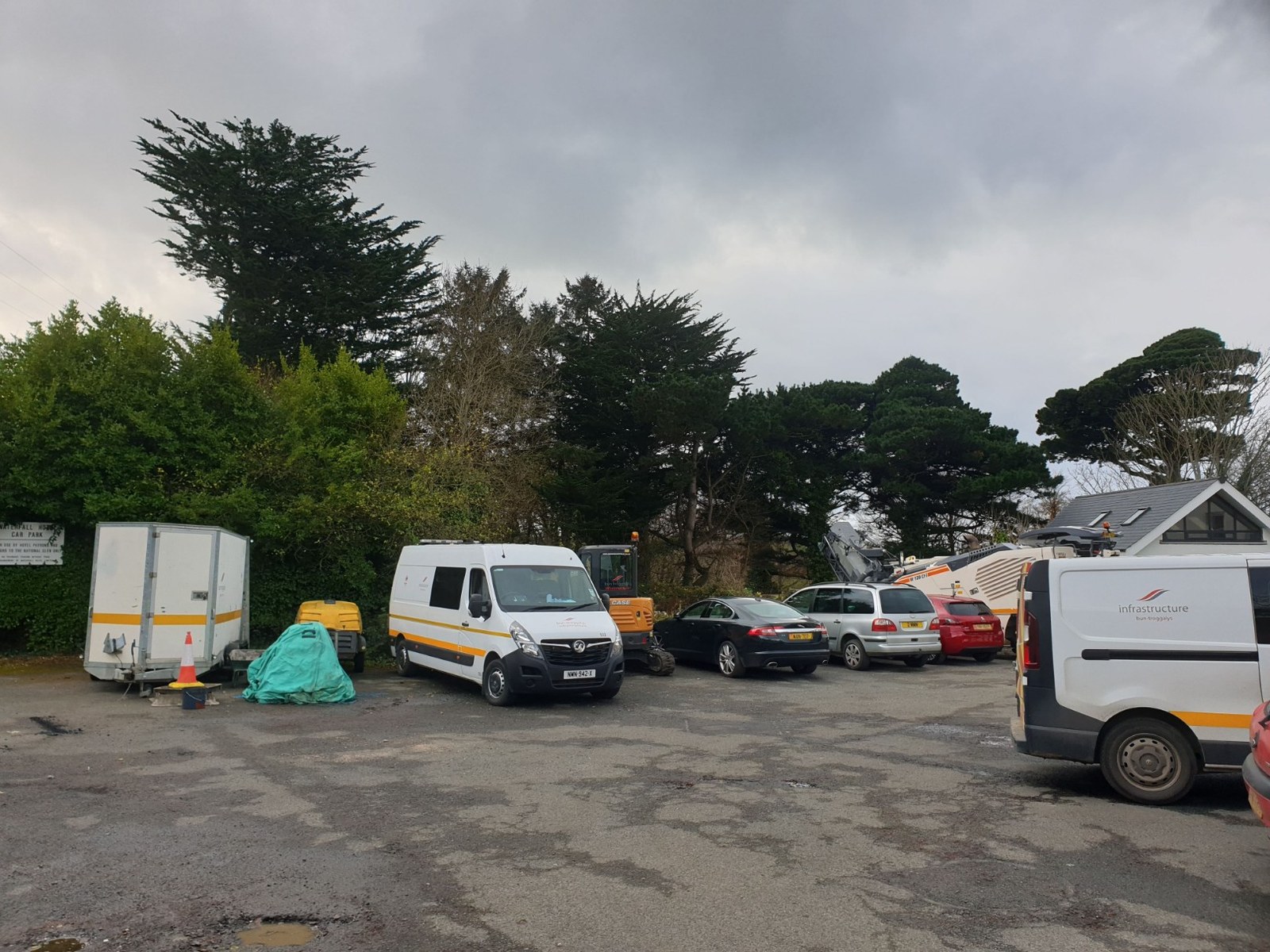 A photograph showing a paved parking area or forecourt with several work vans, cars, and construction equipment parked on it, with trees and a building in the background.