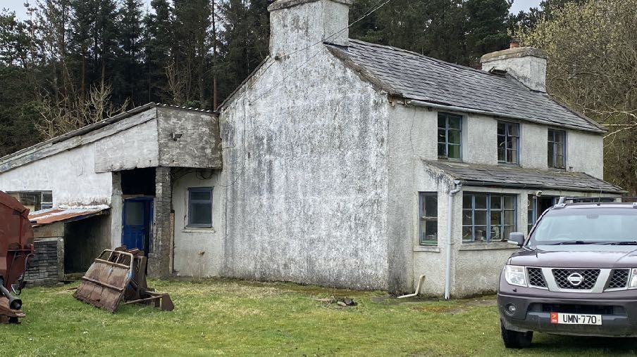A photograph of the existing white two-story dwelling with an attached single-story structure, situated in a rural setting with trees in the background.