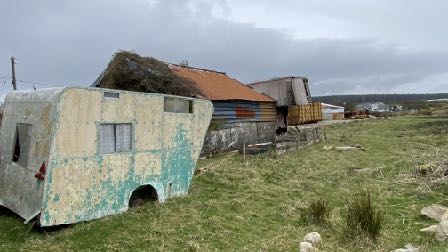 A photograph showing a dilapidated caravan in the foreground with a thatched-roof building and other structures in the background.