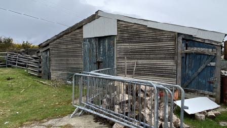 A photograph showing a dilapidated wooden outbuilding or barn with weathered cladding and blue doors, situated in a rural setting with a metal barrier in the foreground.